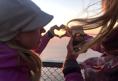 Two women take a photo of their hands making a heart shape at sunset