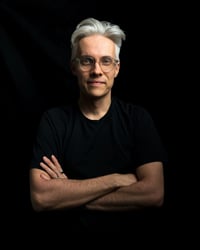 A portrait picture of Lukas Lundin with his hands crossed against a black background. He is wearing a black t-shirt and transparent eyeglasses.