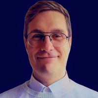 A portrait of Henri Wenlin, Expert at KONE Corporation, wearing black eyeglasses and a white pullover over a dress shirt against a navy blue background.